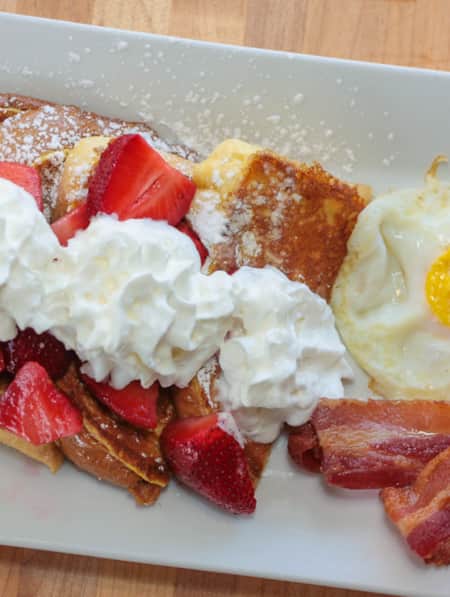 Aerial view of a plate with french toast topped with whipped cream and strawberries, with a fried egg and bacon on the side.