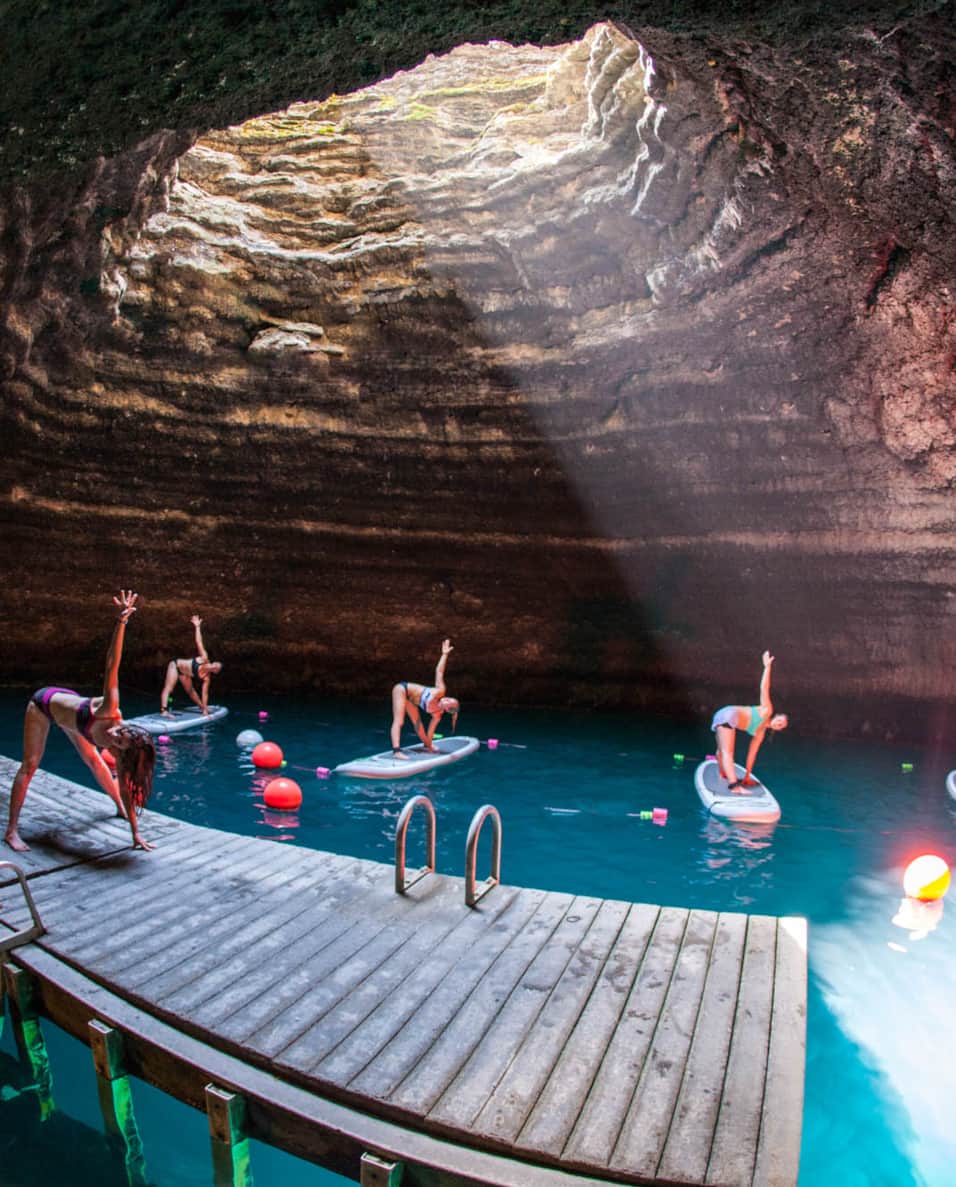 Four women on paddle boards in water doing yoga poses, following an instructor on a wooden dock, all within an underground crater.