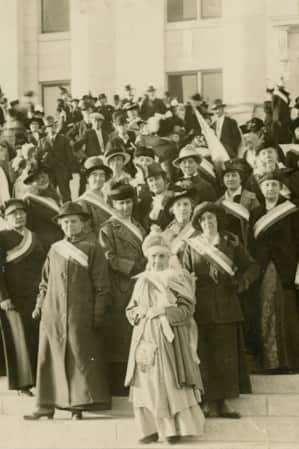 A group of women in long dresses with sashes across their chests, celebrating an event at a government building.