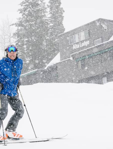 A skier smiles in front of a ski lodge while it is snowing.