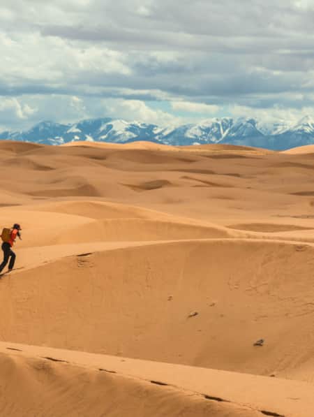 woman hiking through sand dunes with snow-capped mountains in the background.