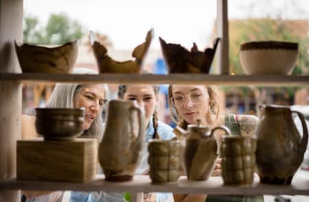 Three women looking at shelves of pottery through a window.