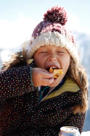 A young girl sitting outside in a ski jacket and hat, taking a big bite of her food at a mountain restaurant.
