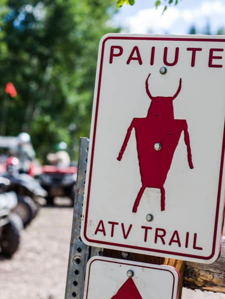 A trail sign reading 'Paiute Trail ATV' with ATVs visible in the background.