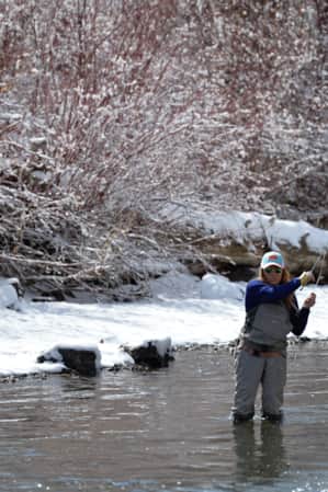 A woman in fly fishing gear standing in a river, with a snow-covered ground along the riverbank