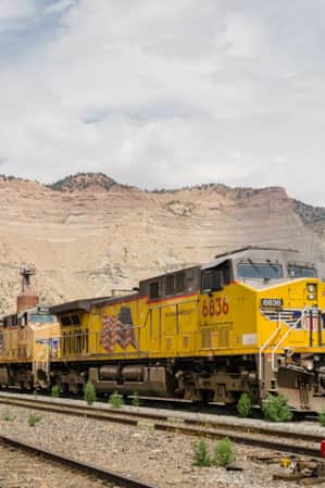 A yellow train arriving in a small town with a large cliff face in the backdrop.