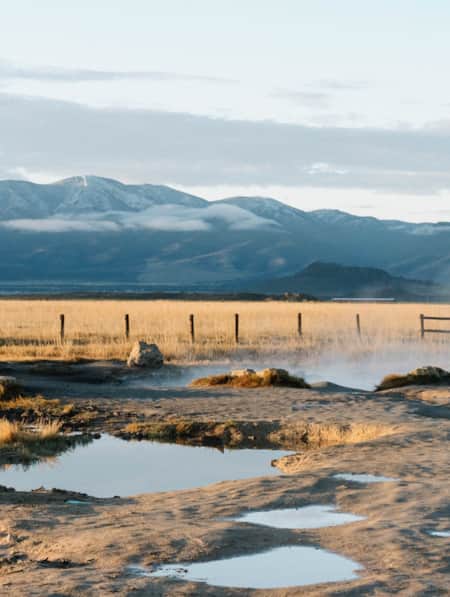 A natural hot spring in a vast field of grass and sand, with a mountain range visible in the distance.