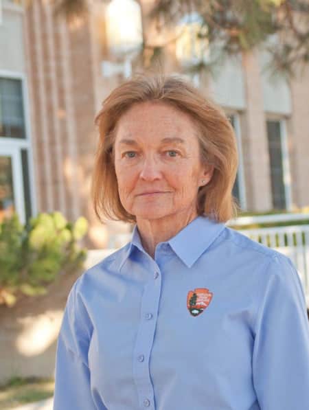 A woman standing in front of a brick building, wearing a blue shirt with the National Park Service logo.