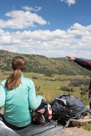 A woman sitting on an ATV looks on as a man standing beside her points toward something in the distance across a vast green valley.