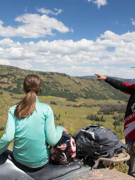 A woman sitting on an ATV looks on as a man standing beside her points toward something in the distance across a vast green valley.
