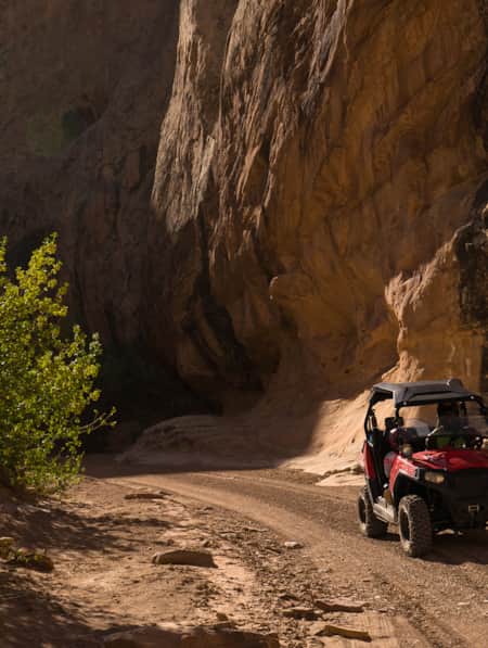 A person riding an ATV along a sandy trail, with greenery on one side and a large rock formation providing shade on the other.