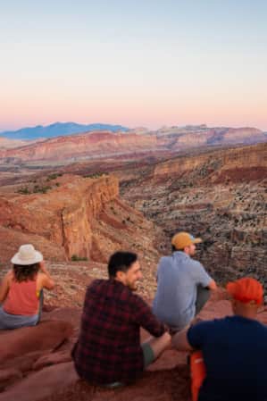 The view from behind of people sitting on a red rock, overlooking a vast canyon that displays multiple layers of rock.