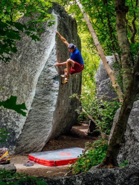 A man climbing a boulder in a forest, surrounded by trees, with a crash pad placed beneath him for a safe landing.