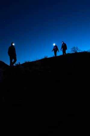 The silhouettes of three hikers with headlamps, standing on the ridge of a hill.