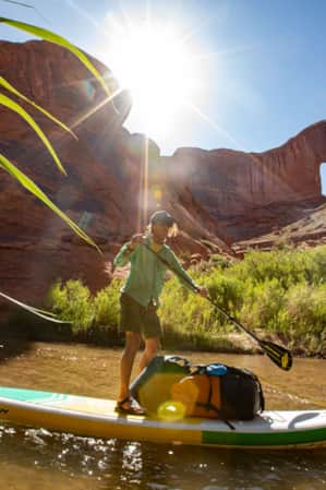 A man stand-up paddleboarding on a river flowing through a red rock canyon during the day.