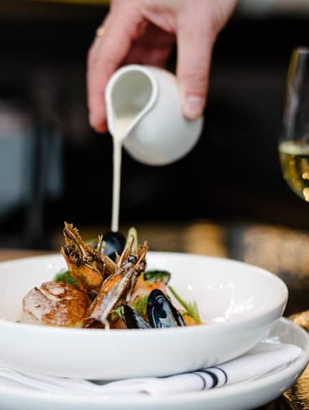 A bowl of elegantly displayed seafood, being topped with a white sauce.