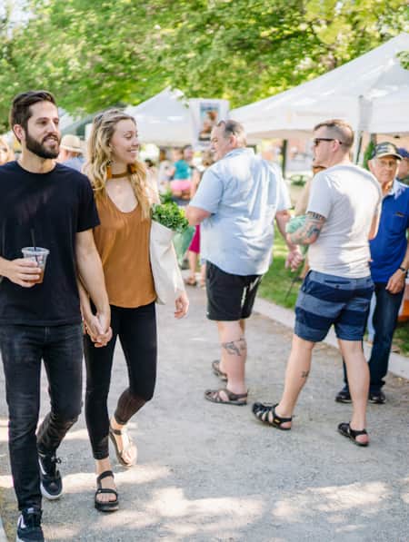 People with bags of fresh produce walking down a gravel path with market vendors lining each side.
