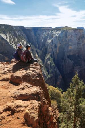 Two people sitting on the edge of a cliff, overlooking a vast red rock canyon with a river winding through it and trees scattered at the canyon's base.