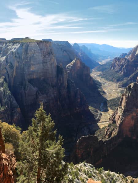 Two people sitting on the edge of a cliff, overlooking a vast red rock canyon with a river winding through it and trees scattered at the canyon's base.