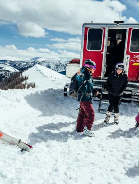A group of people geared up to ski at the top of a mountain, being dropped off by a snowcat.