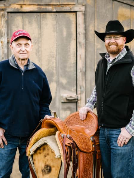 Two men standing on either side of a horse's saddle in front of a barn.