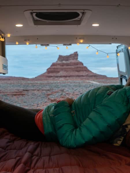 A woman warmly dressed, sitting in a van with a bed set up in the back, gazing out at a large rock formation in the desert.