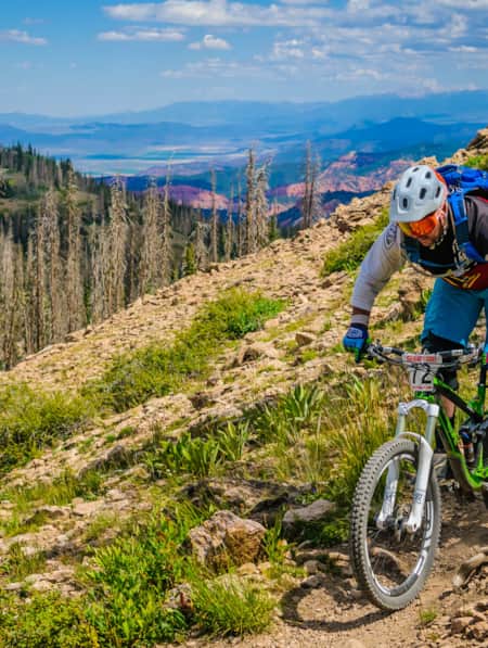 A man mountain biking on a dirt path along the side of a mountain, with additional mountain ranges in the backdrop.