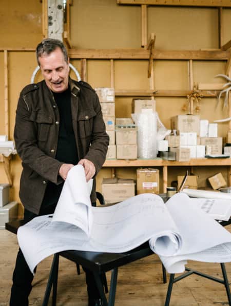A man standing in a warehouse, thumbing through large pieces of paper with building layout plans.
