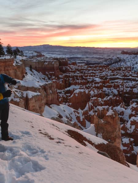 Two hikers overlooking a snowy valley of hoodoos, pouring a warm drink from a thermos.