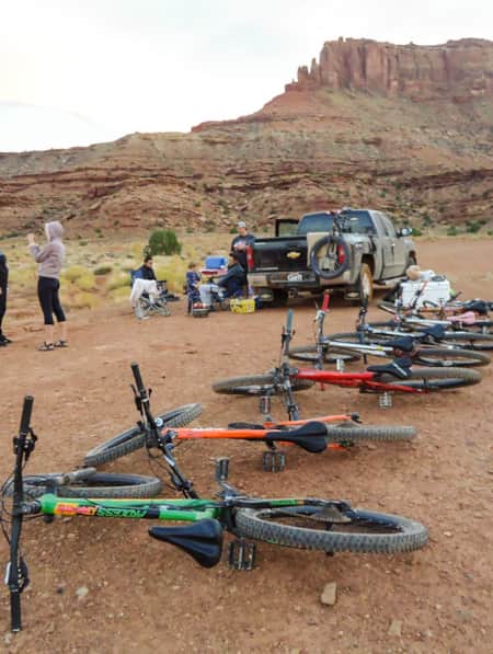 Mountain bikes lying on their sides in a row in the desert, with a group of people by a truck with camping gear off to the side.