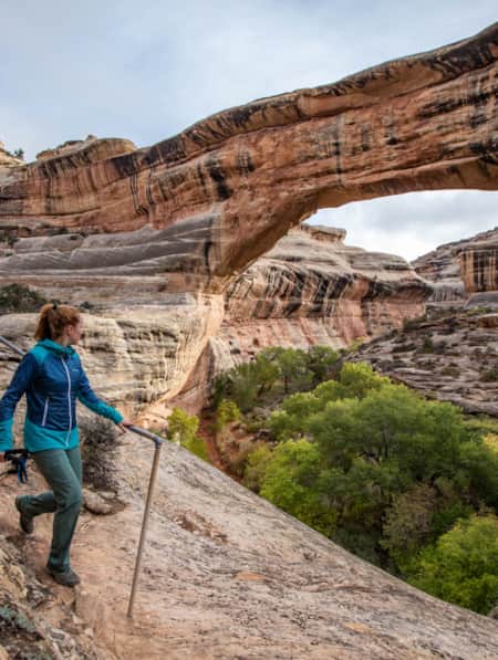 A woman hiking among red rock formations, with a natural bridge visible in the background.
