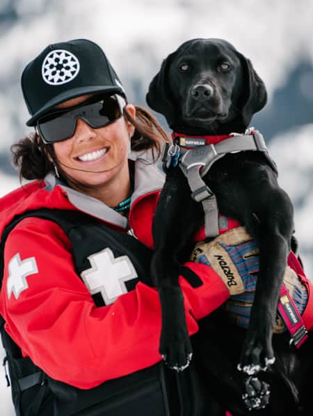 A ski patrol employee smiling while holding a black Labrador Retriever.