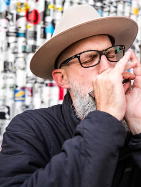 A man playing the harmonica in front of a metal wall covered in stickers.