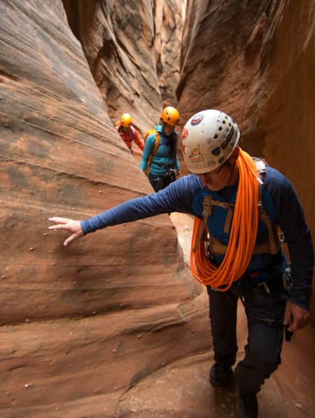 Three people walking through a narrow slot canyon, wearing helmets and carrying rope.