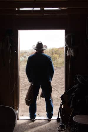 A man standing at the entrance of a barn, wearing chaps and a cowboy hat.
