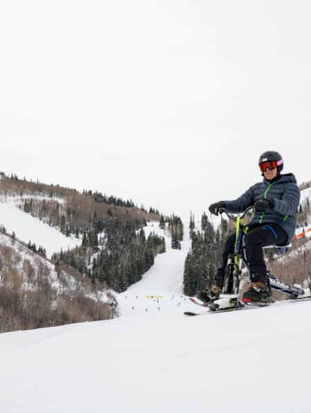 Two people skiing down a mountain: one on special assisted skis, with a man attached by a rope riding behind.