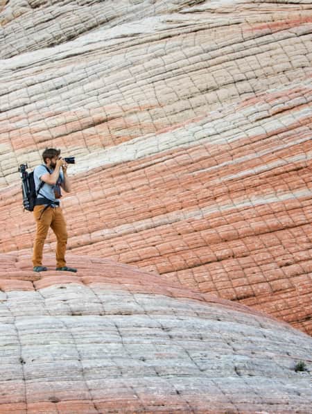 A man with a large camera taking a photo of something in the distance, standing on and surrounded by large rock formations with white and red striations.