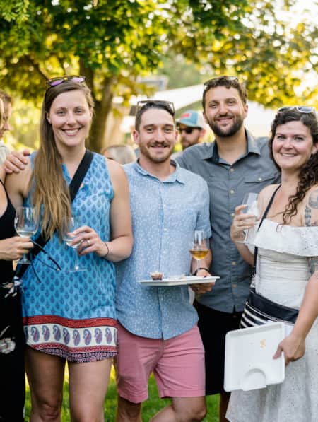 People standing next to each other, all smiling, with some holding wine glasses, at a festival, with others walking around in the background.
