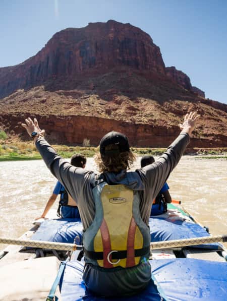 Three people on a large raft in a river, with one person holding their hands in the air, passing by a red rock monument.