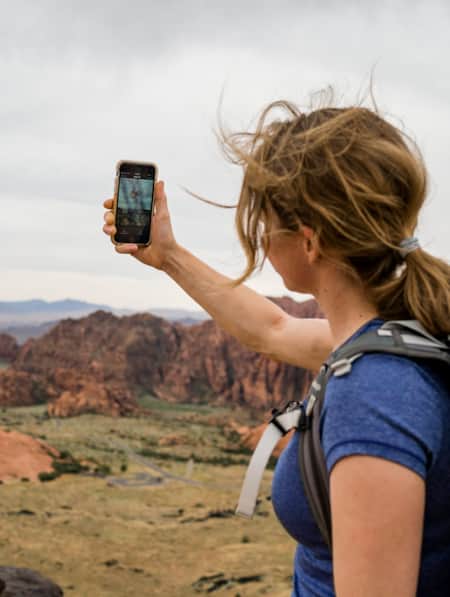 A woman hiking with a backpack, holding up her phone to take a picture of a landscape with sparse vegetation and rock formations.
