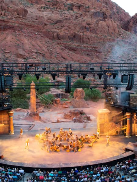An outdoor amphitheater set against a large red rock, with a crowd of people sitting down and watching a performance.
