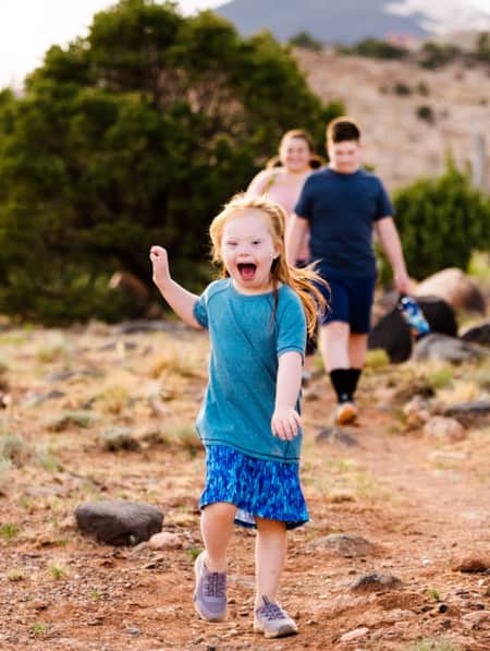 A young girl running on a hiking trail, smiling with her hand up in the air, while her family walks behind her.