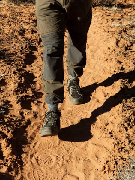 A person's legs walking along a sandy trail with sand on each side covered by a thin layer of a black natural substance.