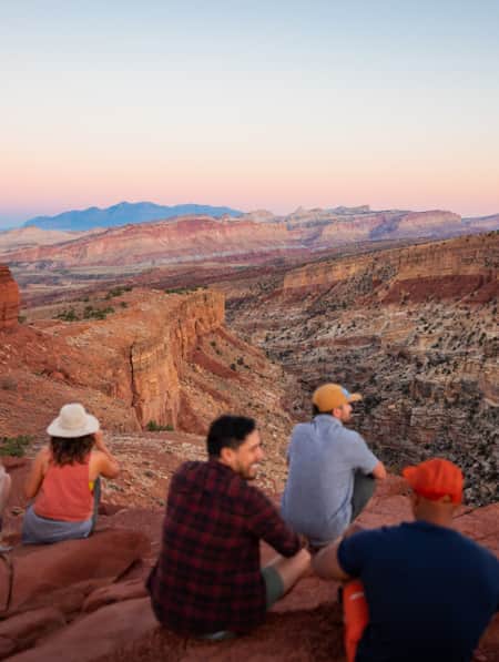 The view from behind of people sitting on a red rock, overlooking a vast canyon that displays multiple layers of rock.