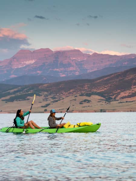 Two women kayaking on a lake, with mountains illuminated pink by the setting sun in the background.