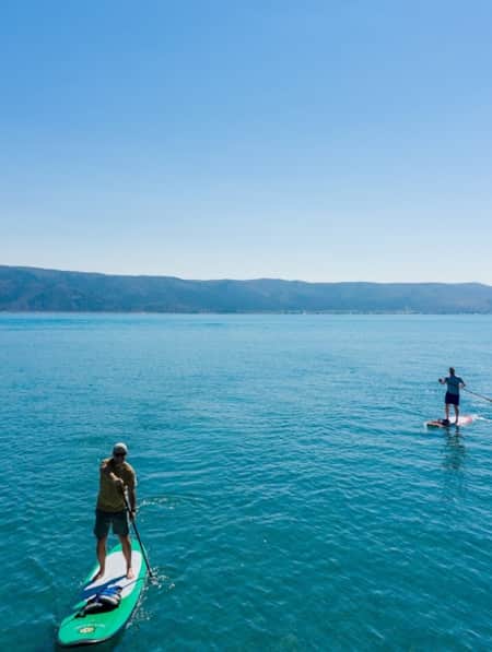 Two men stand-up paddleboarding on a lake with mountains in the backdrop.