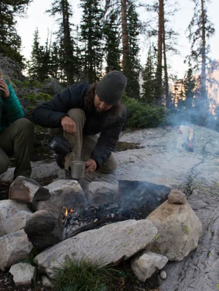 A man and a woman drinking warm beverages around a fire pit in a forest.