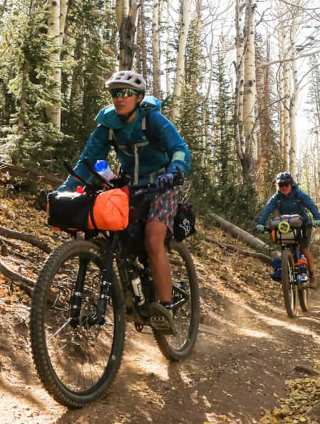 Three people riding mountain bikes in a line, coming down a trail surrounded by trees.