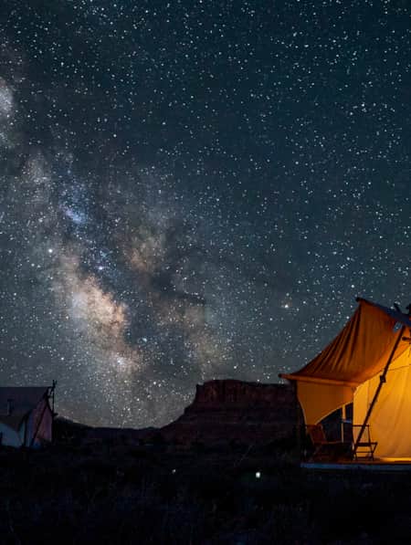 A canvas tent illuminated by a porch light under a starry sky with the Milky Way visible.