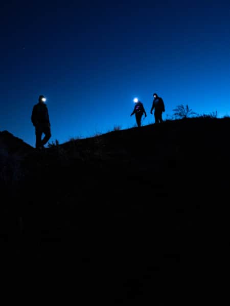The silhouettes of three hikers with headlamps, standing on the ridge of a hill.